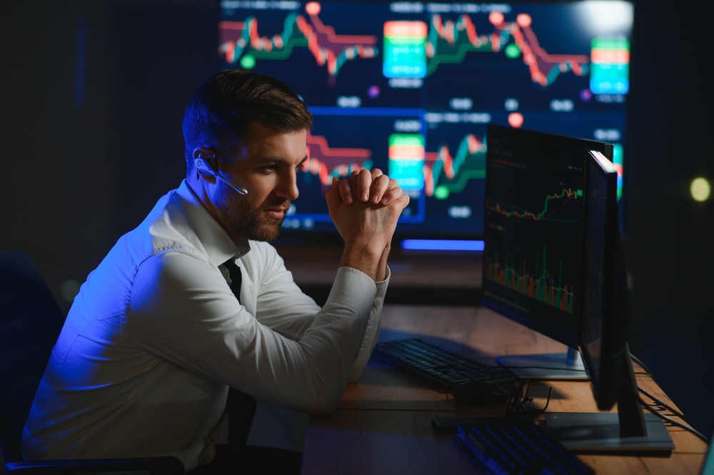 A man wearing a headset sits at a desk, analyzing accounting data and financial charts on his monitors in a dark office with large screens in the background.
