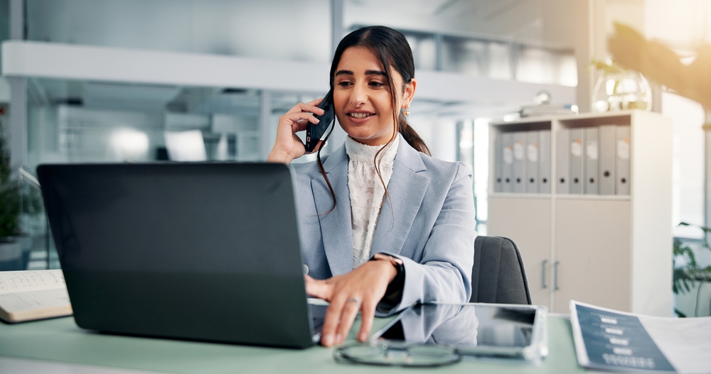 Woman in business attire sits at a desk using a laptop and talking on the phone, demonstrating her Applied Management skills in a modern office setting.