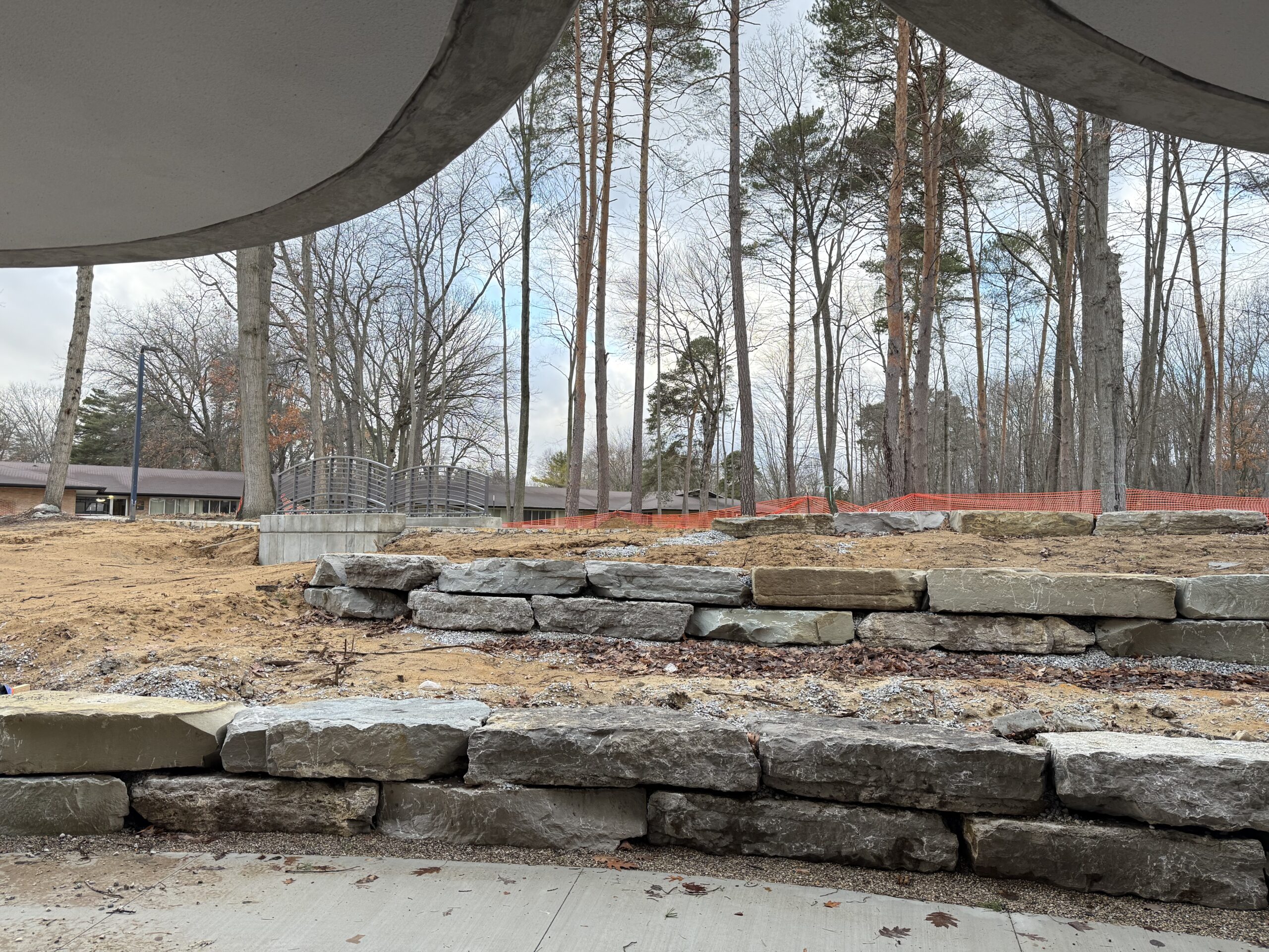 A view of terraced stone retaining walls and bare soil, with trees and orange safety fencing in the background, seen from beneath a concrete overhang.