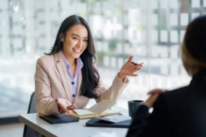 A woman in business attire sits at a desk with an open accounting notebook, gesturing and smiling while talking to another person in an office setting.