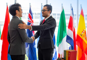 Two men in suits shake hands and smile, standing in front of international flags in an office setting, celebrating a successful economics partnership.