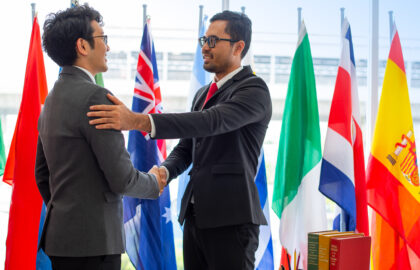 Two men in suits shake hands and smile, standing in front of international flags in an office setting, celebrating a successful economics partnership.