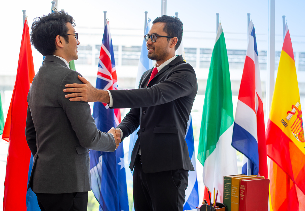 Two men in suits shake hands and smile, standing in front of international flags in an office setting, celebrating a successful economics partnership.