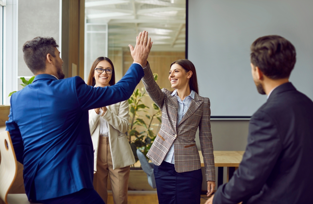 Two people in business attire high-five in an office setting, celebrating an Applied Management success, while two colleagues look on and smile. A presentation screen is visible in the background.