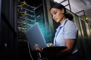 A woman stands in a server room, holding a laptop and working with computer science code, with server racks and cables visible in the background.