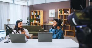Two people wearing headphones and speaking into microphones sit at a table with laptops, recording an entrepreneurship podcast in a modern, well-lit studio with bookshelves and a sofa in the background.