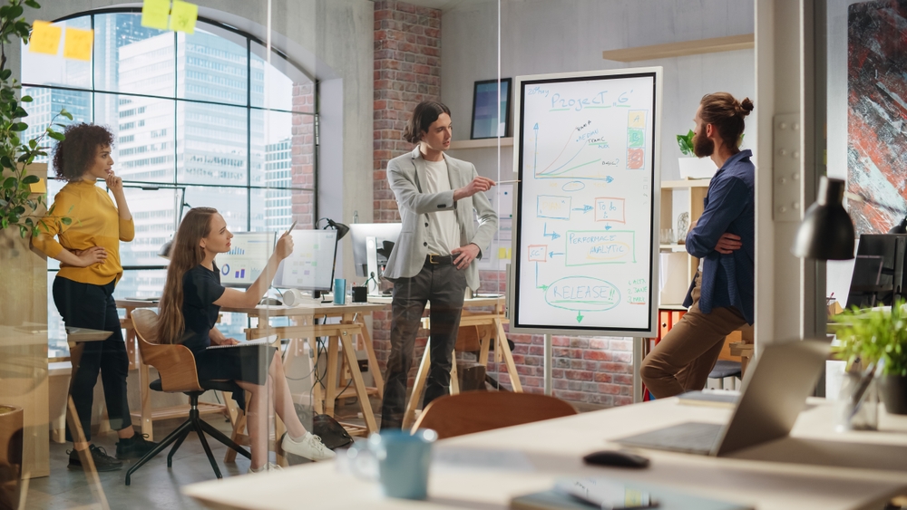 Four people in a modern office discuss a merchandising and marketing project, standing and sitting around a whiteboard filled with charts and diagrams.