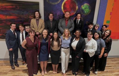 A group of twenty people dressed in business attire pose together indoors in front of colorful abstract artwork, celebrating Northwood University Mock Trial’s participation in the 2025–26 season and the Michigan State Invitational.