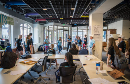 A group of people stands in a brightly lit office space, listening to a speaker near the entrance, while others sit at tables with papers and bags.