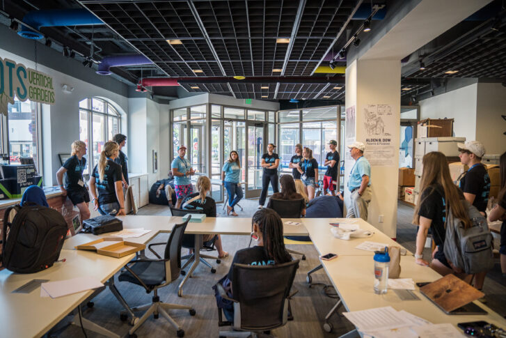 A group of people stands in a brightly lit office space, listening to a speaker near the entrance, while others sit at tables with papers and bags.