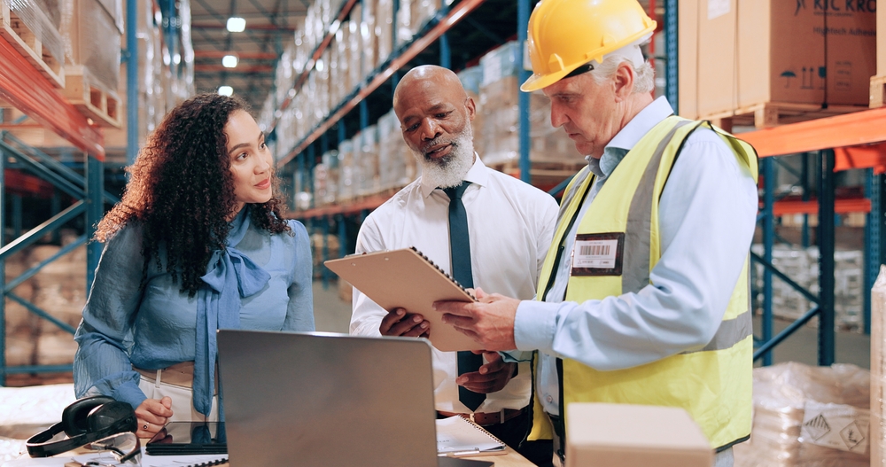 Three people in a warehouse review documents together; one wears a hard hat and safety vest, while the others stand beside a laptop displaying Data Analytics. Shelves with boxes line the background.
