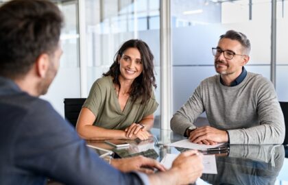 Three people sit at a glass table in an office, engaged in an Automotive Aftermarket Management discussion. Two are facing the camera, smiling, while the third has their back turned. Papers and a phone are on the table.