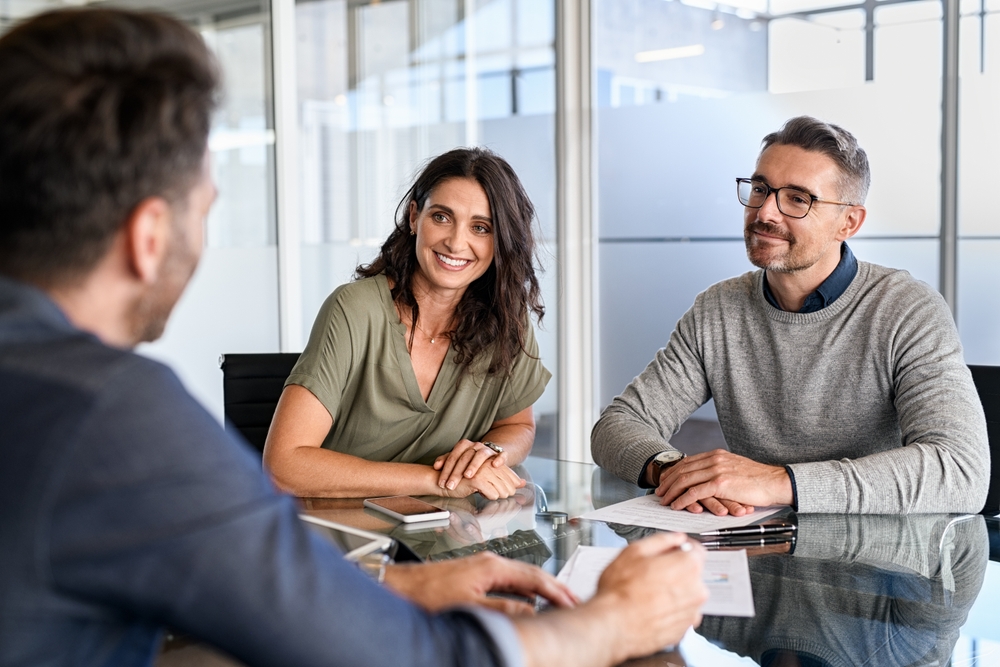 Three people sit at a glass table in an office, engaged in an Automotive Aftermarket Management discussion. Two are facing the camera, smiling, while the third has their back turned. Papers and a phone are on the table.