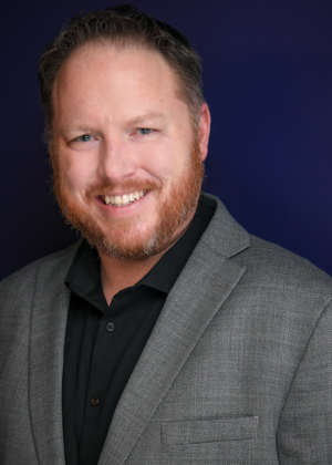 A man with short hair and a beard, wearing a gray blazer over a black shirt, smiles against a dark blue background.