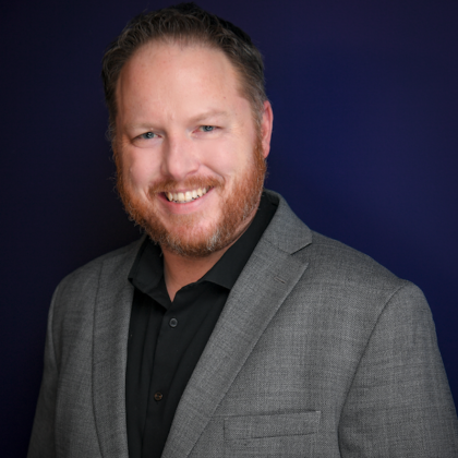 A man with short hair and a beard, wearing a gray blazer over a black shirt, smiles against a dark blue background.