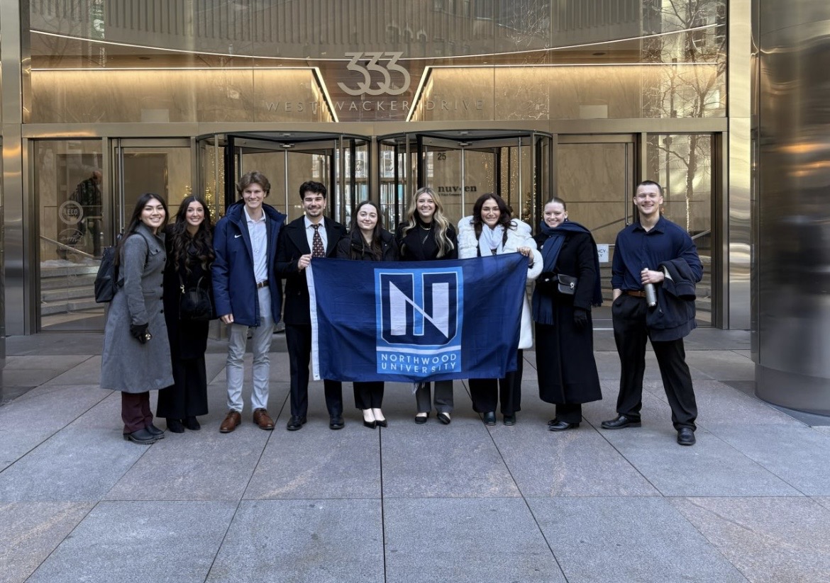 A group of ten people stand outside, holding a blue Northwood University banner and posing for a photo, celebrating their achievements in finance.