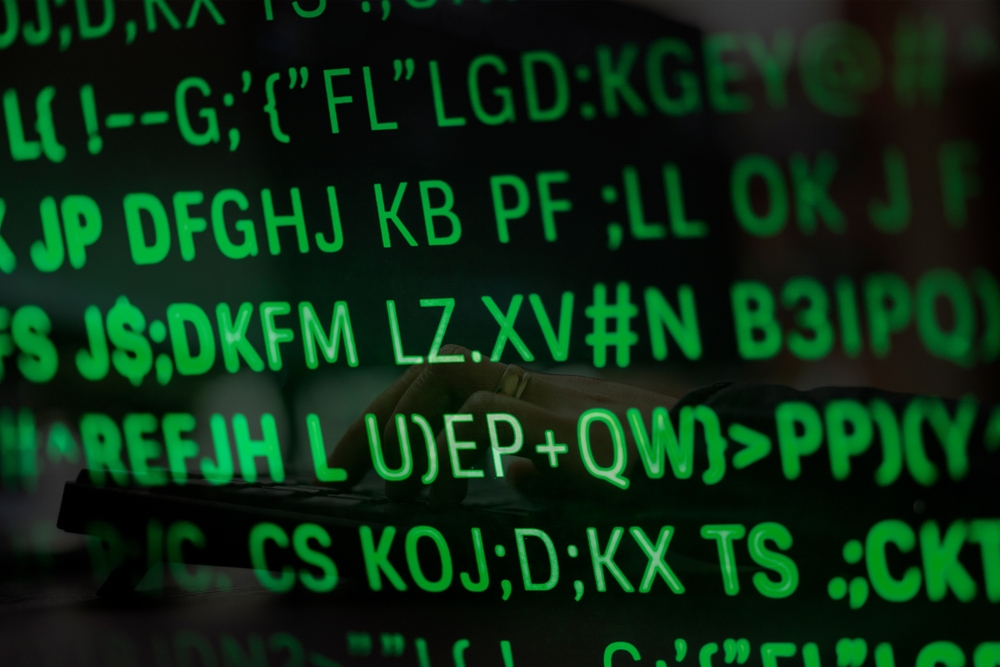 Green alphanumeric code is displayed on a screen, hinting at complex information systems and cybersecurity, with a person's hands typing on a keyboard faintly visible in the background.