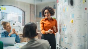 A woman stands at a whiteboard covered with diagrams and sticky notes, presenting concepts of Applied Management to two colleagues seated at a table in a modern office.