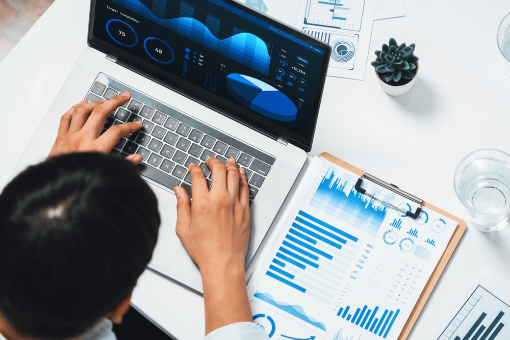 Person working on a laptop displaying finance data visualizations, with printed charts and a clipboard on the desk beside a glass of water and a small potted plant.