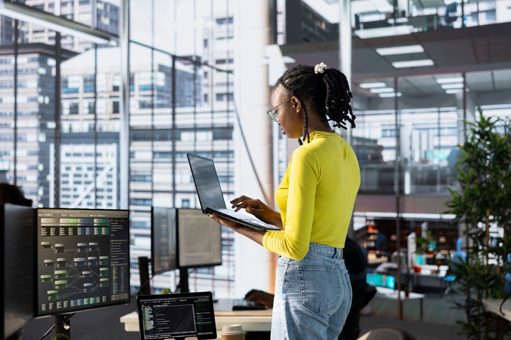 A woman in a yellow shirt stands at a desk in a modern office, working on a laptop. Computer monitors display information systems and cybersecurity data. Large windows show city buildings outside.