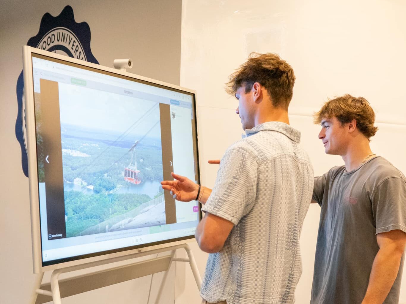 Two young men interact with a large touchscreen display showing an image of a cable car over a landscape in a brightly lit indoor space.