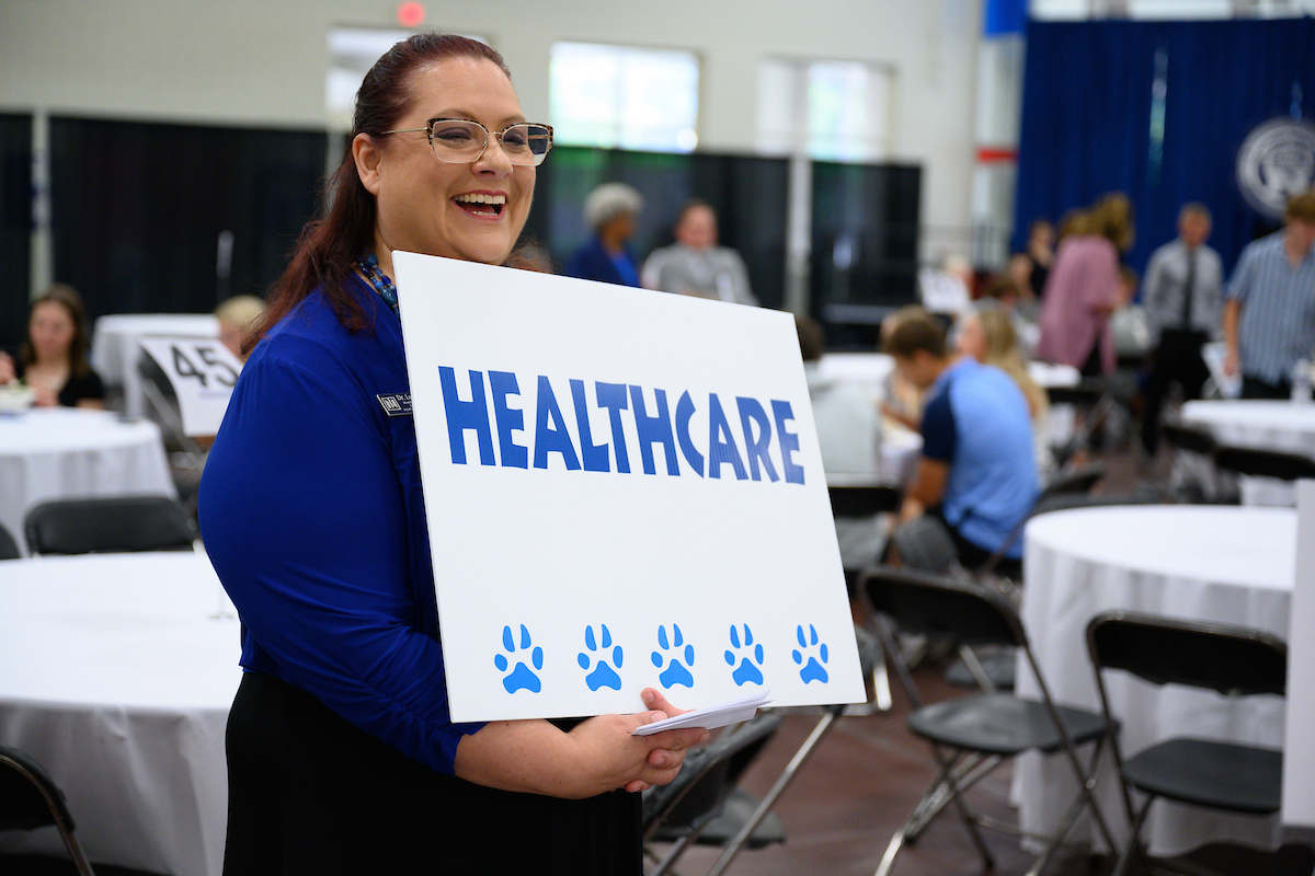 A woman holds a sign reading "HEALTHCARE" with five paw prints below it, highlighting health care management in a room with round tables and several people in the background.