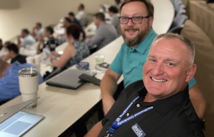 Two men sit at a curved desk in a lecture hall, smiling at the camera. One wears a lanyard and name tag. Other automotive professionals are seated in the background, likely attending University of the Aftermarket 2026 courses.