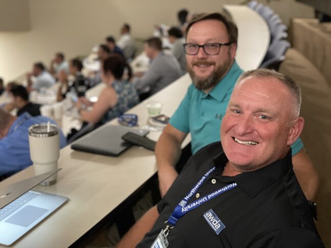 Two men sit at a curved desk in a lecture hall, smiling at the camera. One wears a lanyard and name tag. Other automotive professionals are seated in the background, likely attending University of the Aftermarket 2026 courses.