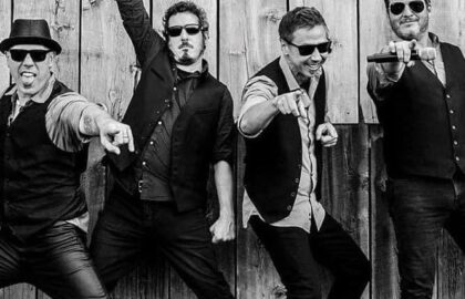 Four men in vests and sunglasses pose enthusiastically in front of a wooden wall, with three pointing at the camera and one raising a fist, capturing a moment that radiates freedom and excitement for the future.