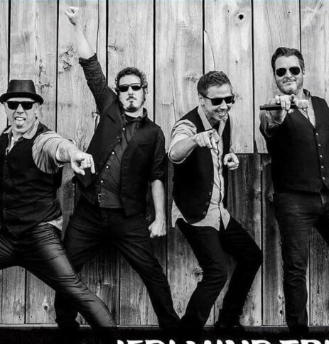 Four men in vests and sunglasses pose enthusiastically in front of a wooden wall, with three pointing at the camera and one raising a fist, capturing a moment that radiates freedom and excitement for the future.