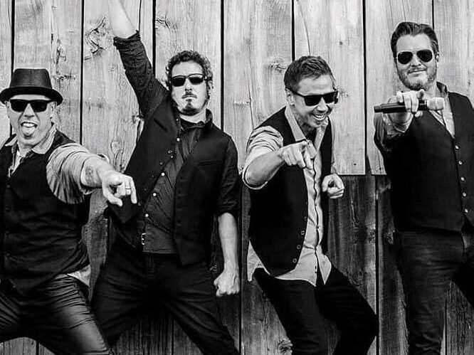 Four men in vests and sunglasses pose enthusiastically in front of a wooden wall, with three pointing at the camera and one raising a fist, capturing a moment that radiates freedom and excitement for the future.