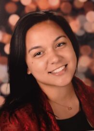 A woman with long dark hair, a 20 Under 40 Alumni, smiles at the camera, wearing a red patterned top and a necklace, with a blurred light background.