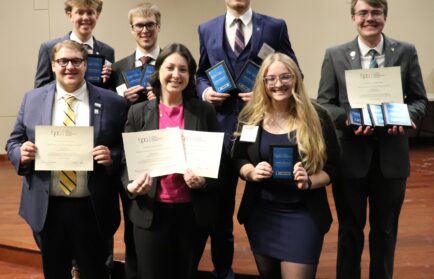 Seven people in business attire stand on a stage, smiling and holding certificates, plaques, and awards.