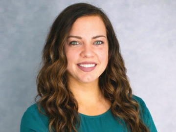 20 Under 40 Alumni, this woman with long wavy brown hair wearing a teal blouse smiles confidently with arms crossed, standing in front of a plain light gray background.
