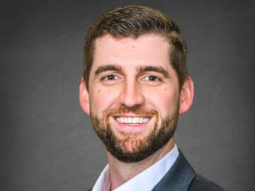 A man with short brown hair and a beard, wearing a dark suit jacket and white shirt, smiles at the camera against a plain dark background, proudly representing the 20 Under 40 Alumni.