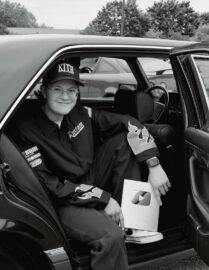 A 20 Under 40 Alumni wearing glasses, a cap, and a racing jacket sits in the backseat of a car, proudly holding a YouTube Silver Play Button award.