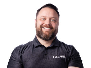 Man with a beard smiling, wearing a black polo shirt that says "LEAD.WIN." on the left chest, standing against a plain white background—an inspiring alumni recognized in the 20 under 40 list.