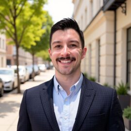 A 20 Under 40 alumni in a suit smiles at the camera while standing on a sunny sidewalk lined with trees and parked cars.