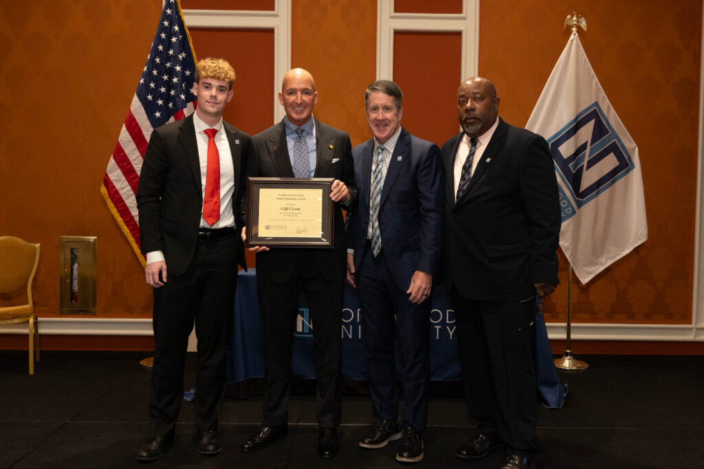Four men in suits stand together; one holds a framed certificate. An American flag and another flag are visible in the background.