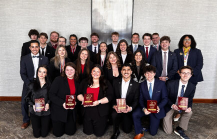 A group of young adults in business attire pose indoors, some kneeling in front and holding awards, with a neutral-colored wall and artwork in the background.