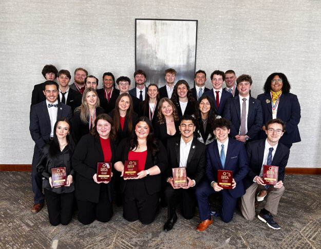 A group of young adults in business attire pose indoors, some kneeling in front and holding awards, with a neutral-colored wall and artwork in the background.