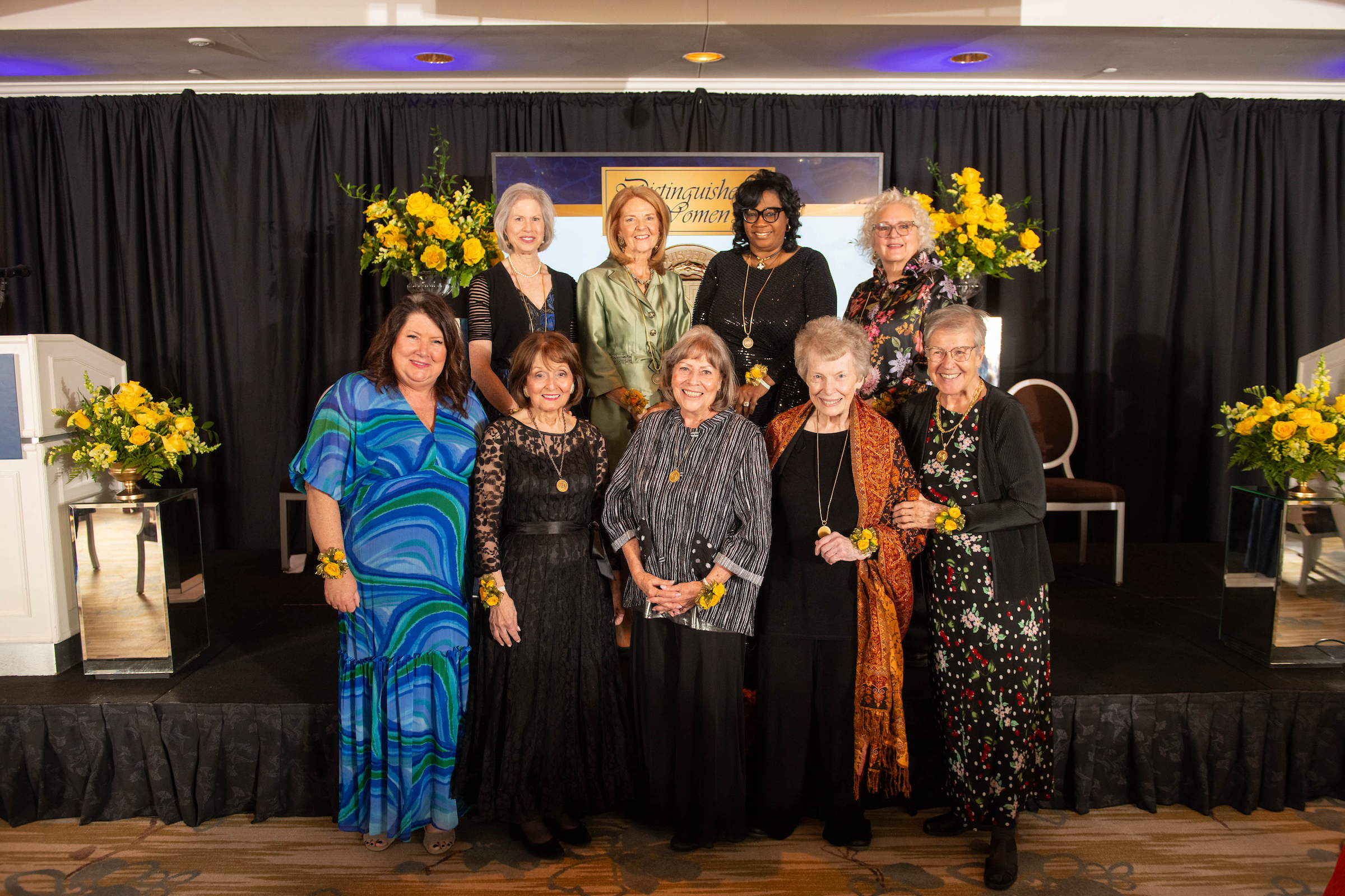 Nine women pose together on a stage with floral arrangements and a banner for the Northwood's Distinguished Women Awards, all dressed in formal attire.