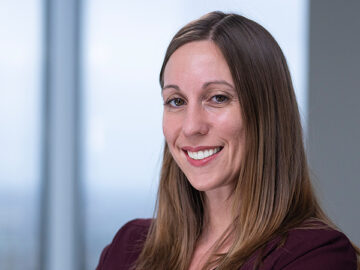 A woman with straight brown hair smiles with arms crossed, wearing a burgundy blazer and black top, standing in a modern office with large windows—a proud Alumni featured in the 20 Under 40 list.