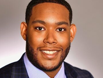 A 20 Under 40 Alumni, dressed in a checked navy suit, light blue shirt, and patterned tie, smiles at the camera against a neutral background.