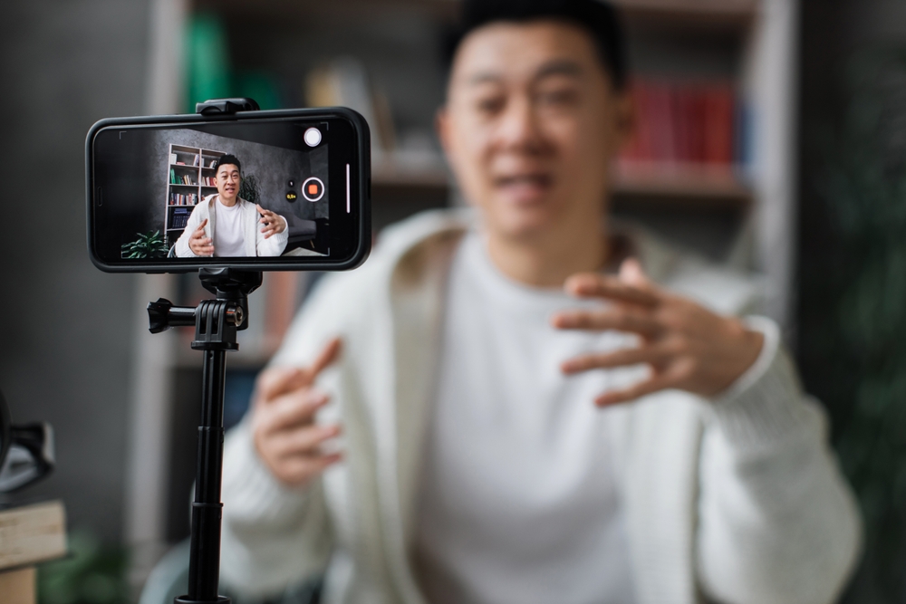A person gestures while speaking in front of a smartphone camera mounted on a tripod, recording a marketing video in an indoor setting with shelves in the background.