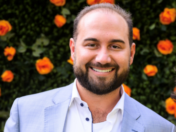 A man in a light gray suit and white shirt, recognized as a 20 Under 40 Alumni, stands in front of a wall decorated with orange roses, smiling at the camera.
