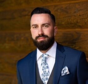 A 20 Under 40 alumni with a dark beard and short hair, wearing a navy blue suit, white shirt, and floral tie, stands in front of a wooden wall.