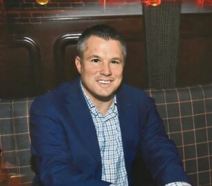 A 20 Under 40 Alumni in a blue suit jacket and checkered shirt sits on a plaid-patterned bench, smiling at the camera in a dimly lit indoor setting.