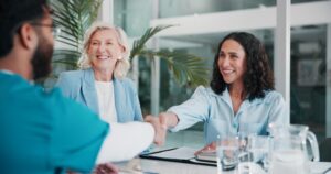 Two women sit at a table, smiling and shaking hands with a man in medical scrubs; documents related to health care management and water pitchers are on the table in a modern office setting.