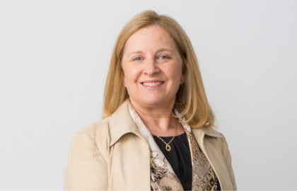 A woman with blonde hair, wearing a beige blazer and patterned blouse, smiles at the camera against a plain light background, embodying confidence and Leadership Insights.